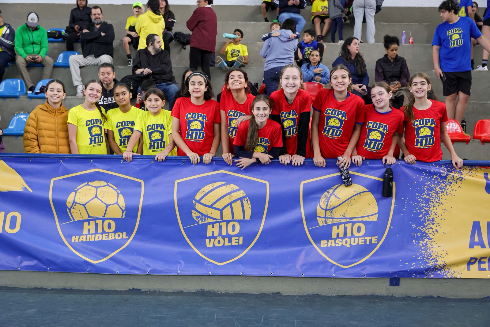 Alunas da H10 e famílias reunidas na arquibancada, apoiadas no banner da Copa H10
